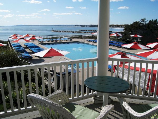 A coastal resort scene with a pool, red and white umbrellas, blue loungers, and a balcony view overlooking the ocean under a clear sky.