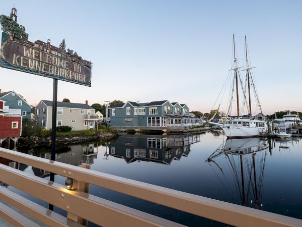 A harbor scene with boats, waterfront buildings, and a "Welcome to Kennebunkport" sign reflecting in calm water.