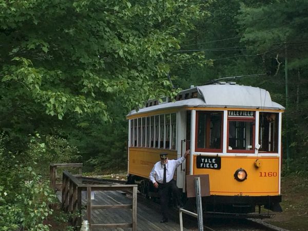 A vintage yellow and white trolley labeled "Yale Field" with a person standing near the door, amidst a forested backdrop.