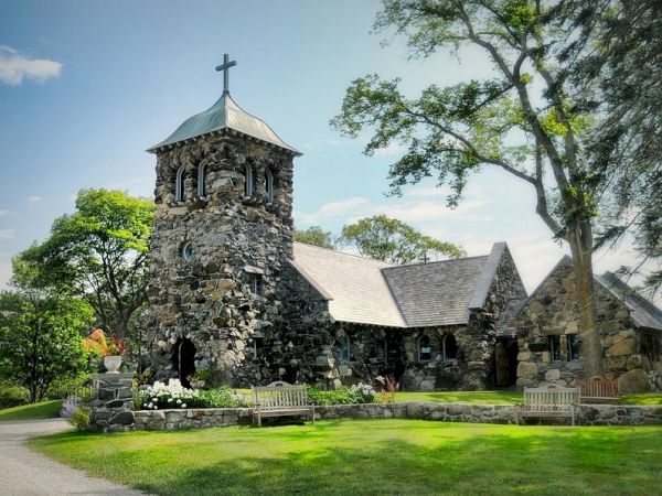 A rustic stone church with a bell tower and cross stands amidst a lush, green landscape with trees and benches in the foreground.
