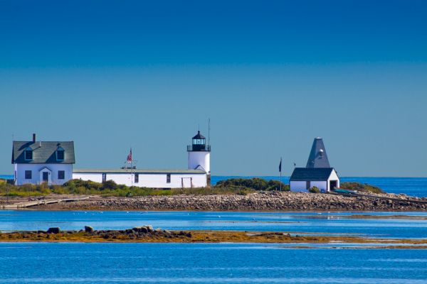 A quaint lighthouse and surrounding buildings sit on a rocky coastal strip, with calm blue waters in the foreground and clear skies above.
