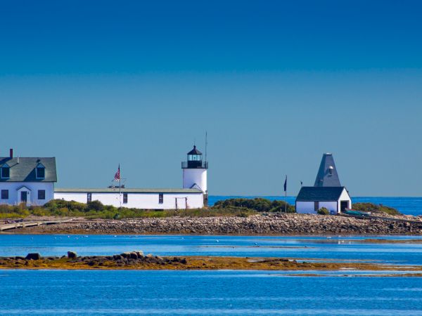 A quaint lighthouse and surrounding buildings sit on a rocky coastal strip, with calm blue waters in the foreground and clear skies above.