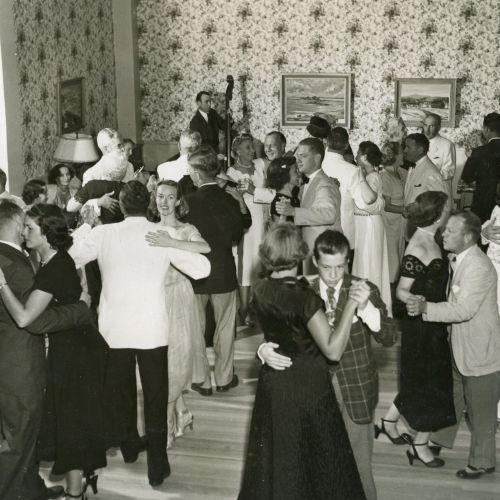 A vintage black-and-white photo shows people dressed formally, dancing in a room with floral wallpaper and paintings on the walls.