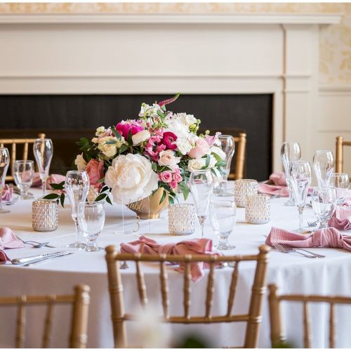 A beautifully set round table with pink napkins, floral centerpiece, glassware, and gold chairs, in an elegant room setting.