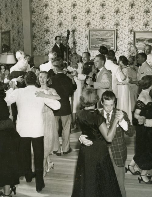 A vintage black and white photo shows a classy indoor dance party with couples dancing, dressed in formal attire, and enjoying the event.