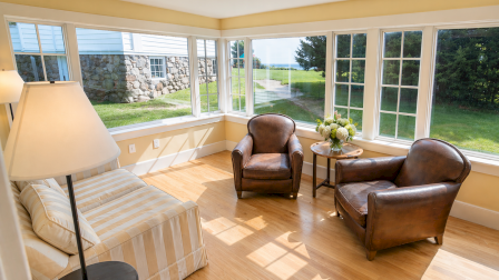 A bright sunlit living room with beige striped sofa, two brown leather chairs, a small side table with flowers, and large windows opening to a green lawn.
