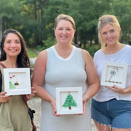 Three people outdoors holding framed art with small tree and nature designs.