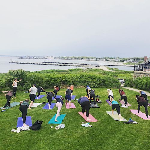A group of people is practicing yoga on colorful mats, outdoors near the sea, on a grassy area with a scenic ocean view.