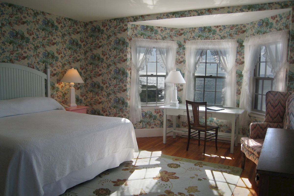A cozy bedroom features floral wallpaper, a large bed, a desk by bay windows, two lamps, and sunlight streaming onto a patterned rug.