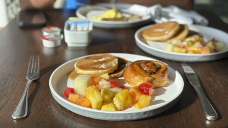 Plates with pancakes, a pastry, and fruit, set on a table with cutlery, and a person in the background.