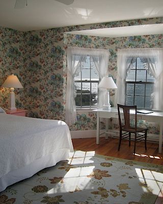 A cozy bedroom with floral wallpaper, a white bedspread, a chair at a window desk, and a sunny rug, in a bright, inviting atmosphere.