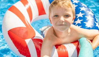 A child is in a pool, holding onto a noodle, and using a red, white, and blue inflatable ring with stars and stripes.