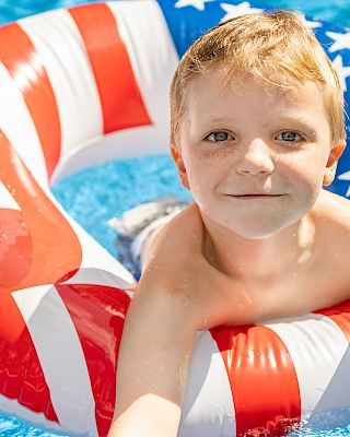 A child is in a pool, floating on an American flag-patterned inflatable tube, smiling, and holding a foam pool noodle.