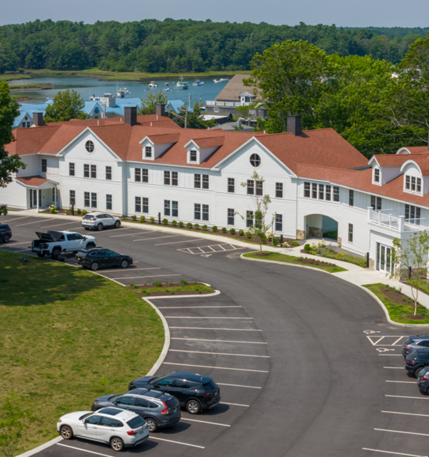 A white building with red roofs, adjacent to a parking lot, is set against a backdrop of trees and a body of water.