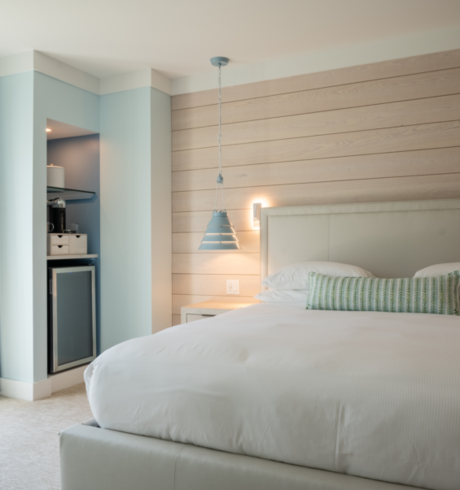 A cozy bedroom with a large bed, modern pendant light, blue curtains, a small kitchenette with a fridge, and a balcony view.