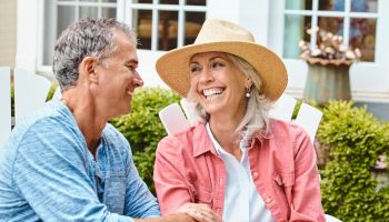 A couple sits on outdoor chairs, smiling at each other. The man wears a blue shirt, the woman a pink shirt and straw hat. Bushes are in the background.