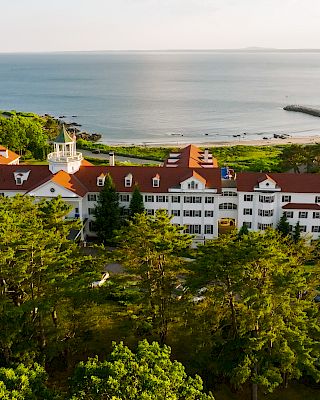 A large white building with a red roof is surrounded by trees, overlooking a coastal area with calm waters and a rocky shoreline.