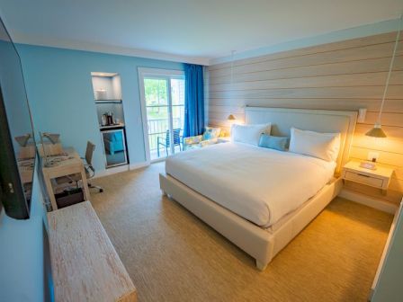 A modern hotel room with a large bed, desk, and balcony. Light decor adds a cozy touch, with natural light filtering through the window.