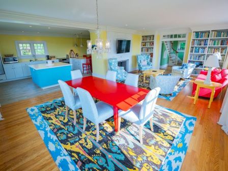A bright, colorful living space with a red dining table, blue kitchen island, bookshelves, and various seating areas on a patterned rug.