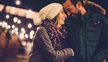A couple bundled up in cozy coats, sharing a warm, smiling moment outdoors at night with twinkling lights in the background.