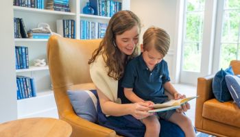 A woman and a child are sitting in a cozy room, reading together on a chair. A bookshelf full of books is in the background.