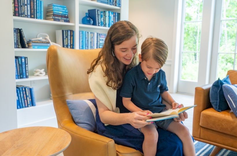 A woman and a child are sitting in a cozy room, reading together on a chair. A bookshelf full of books is in the background.