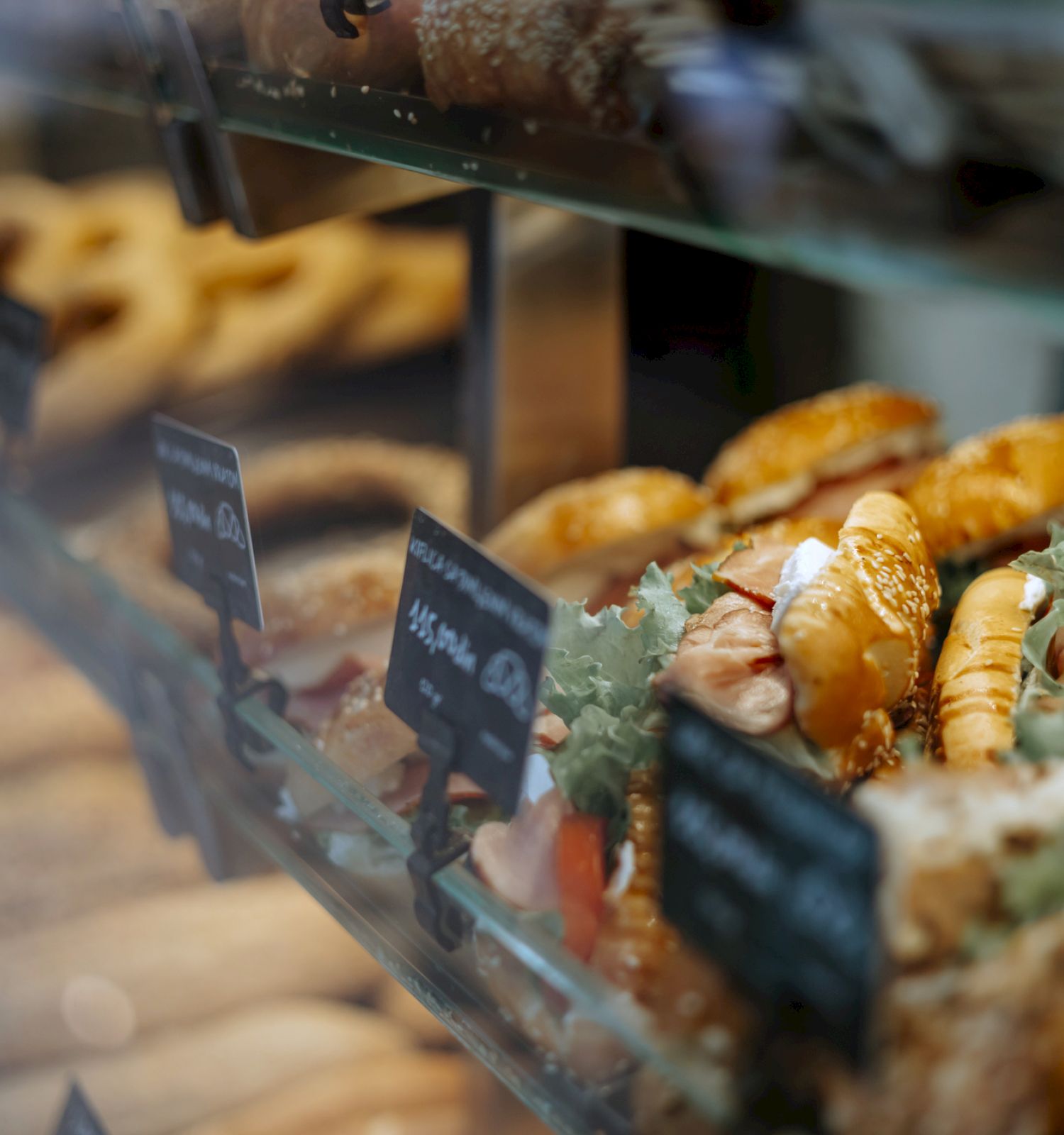 A display case filled with various pastries and sandwiches, some with visible sesame seeds, alongside small signs.