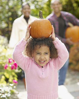 A child holds a pumpkin above their head while smiling, with two adults in the background, surrounded by a garden setting.