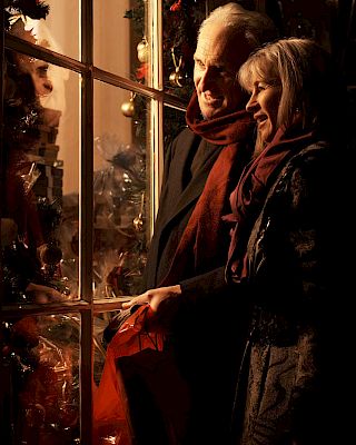 A couple is window shopping, warmly dressed with scarves, admiring a festive display, with Christmas decorations and wrapped gifts inside.
