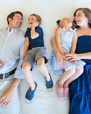 A family of four lying on a bed, smiling and looking at each other, wearing casual clothing.