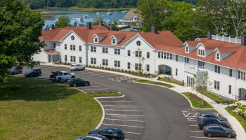 A large building with a red roof, parking lot with cars, and a view of water and trees in the background.