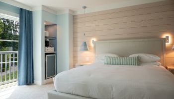 A cozy bedroom with a large bed, soft lighting, blue curtains, and a small kitchenette area with a mini fridge and coffee maker.