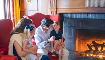 A family sitting by a cozy fireplace in a warmly decorated living room with a painting and vases on the mantel.