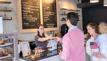 A family is at a cafe counter while a staff member serves them. Menu boards and baked goods are visible in the bright interior.