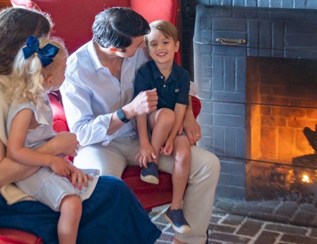 A family of four sits by a lit fireplace on a cozy brick hearth, engaging in a warm and intimate moment together.