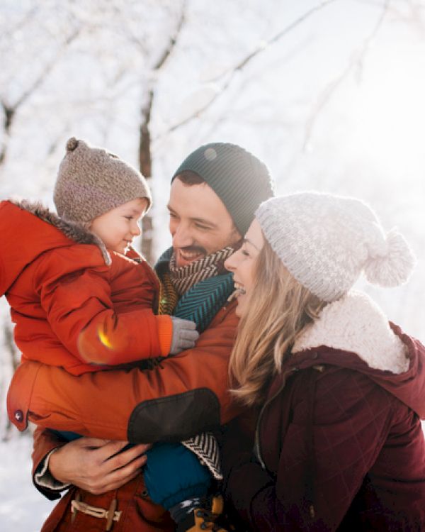 A family of three in a snowy forest: a smiling mom and dad hold their child in warm winter coats and knit hats, sunlight glows softly.