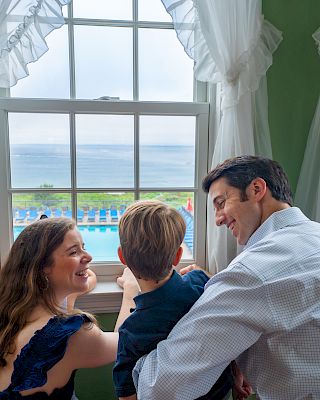 A family of three is looking out a window with a view of a pool and the ocean in the background, framed by white curtains.