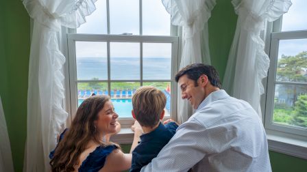 A family of three is looking out a window with a view of a pool and the ocean in the background, framed by white curtains.