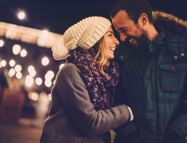 Two people are smiling and embracing on a cozy night street, warmly dressed, enjoying a festive, candle-lit atmosphere.