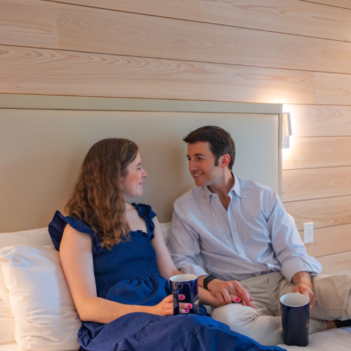 A couple sits on a bed, smiling and talking, enjoying drinks together in a cozy, wooden-paneled hotel room.