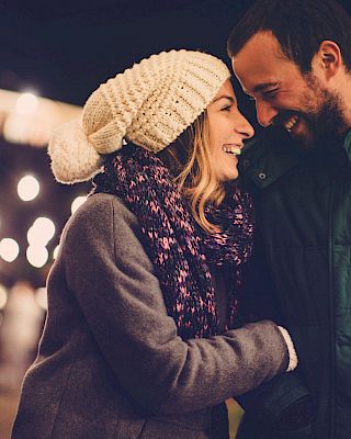 A couple smiling and cuddling in winter coats and scarves at night, glowing lights in the background, warm and romantic mood.